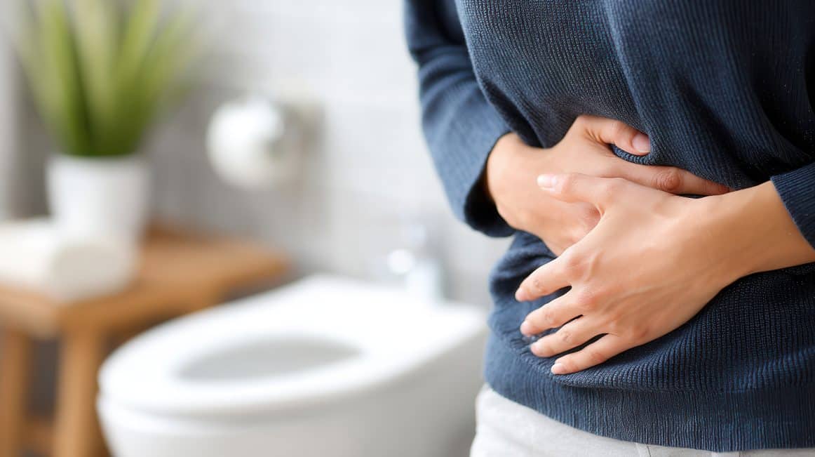 Frau im Vordergrund, die sich den Bauch hält. Im Hintergrund ist eine Toilette zu sehen