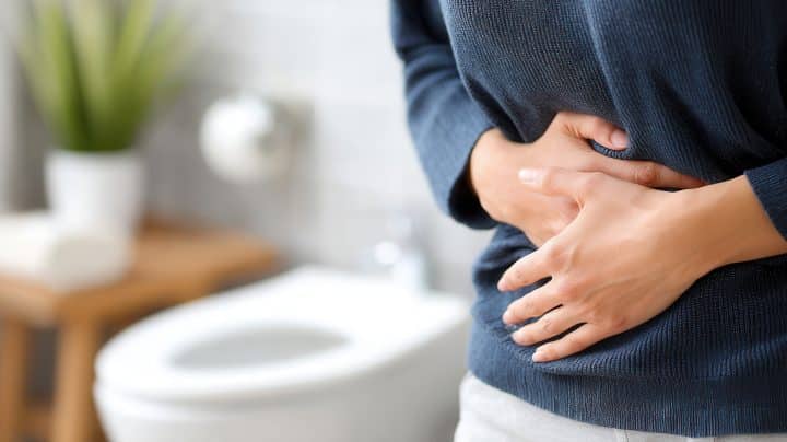 Frau im Vordergrund, die sich den Bauch hält. Im Hintergrund ist eine Toilette zu sehen