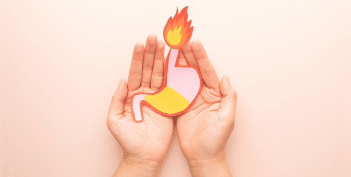 Hände, die einen Magen aus Papier hält. Es ist die Magensäure zu sehen und eine Flamme, die aus der Speiseröhre kommt um Sodbrennen darzustellen