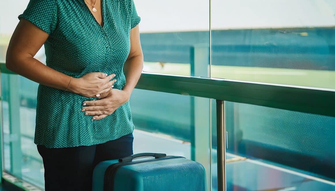 Eine Frau steht mit Koffer am Gate im Flughafen und hält sich den Bauch mit Magenschmerzen