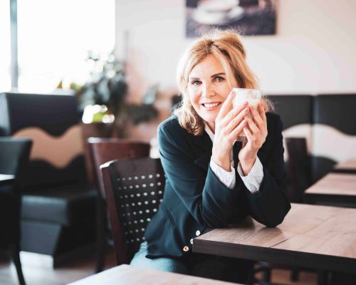 Attraktive,  ältere Frau sitzt im Cafe, hält einen Latte Macchiato in der Hand und lächelt freundlich in die Kamera