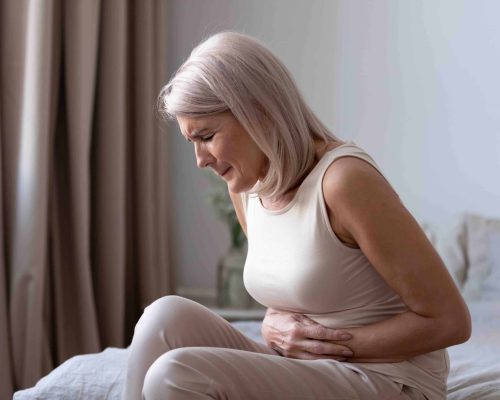 Middle-aged woman sitting on bed feels unhealthy touch stomach suffers from severe crampy abdominal pain, caused by indigestion or diarrhoea, gastric ulcer gastritis problem or unhealthy diet concept